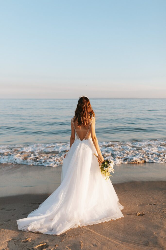 Femme en robe de mariée sur une plage face à la mer et qui tient un bouquet de fleurs